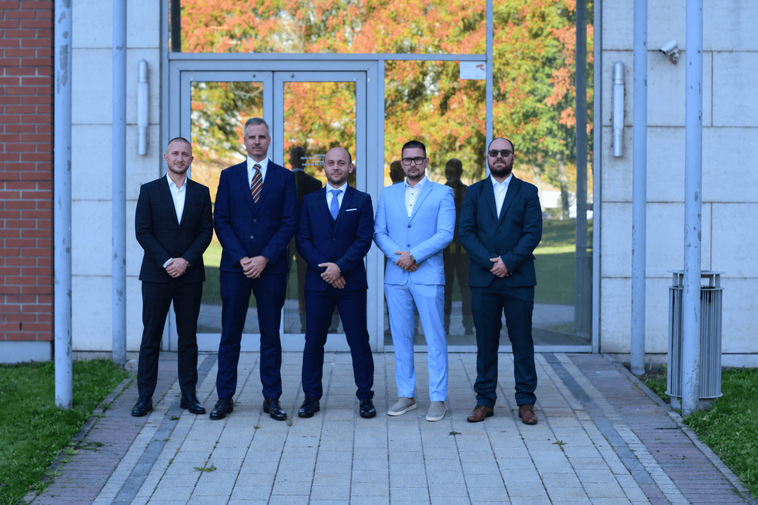 Five men in suits outside a building entrance in an autumn setting